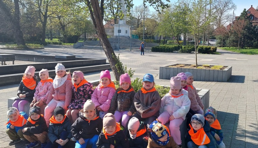 Grupa małych dzieci w pomarańczowych kamizelkach siedzi w Parku Słowiańskim w Głogowie. W tle drzewa, zabudowania i plac