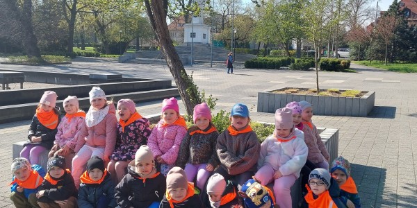 Grupa małych dzieci w pomarańczowych kamizelkach siedzi w Parku Słowiańskim w Głogowie. W tle drzewa, zabudowania i plac