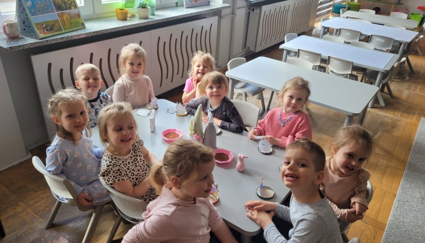 Grupa jedenastej dzieci siedząca przy białych stołach w jasnej sali lekcyjnej, uśmiechająca się do kamery podczas przygotowań