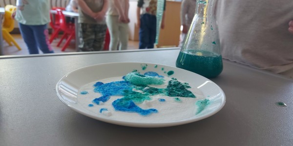 Biały talerz z niebiesko-zielonymi plamami barwnika, obok szklanka z zielonym płynem.