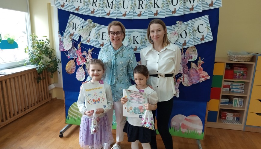 Dwie dorosłe kobiety i dwie małe dziewczyny pozują przed dekoracją z napisem Marmurkowa Wielkanoc w przedszkolnej sali.