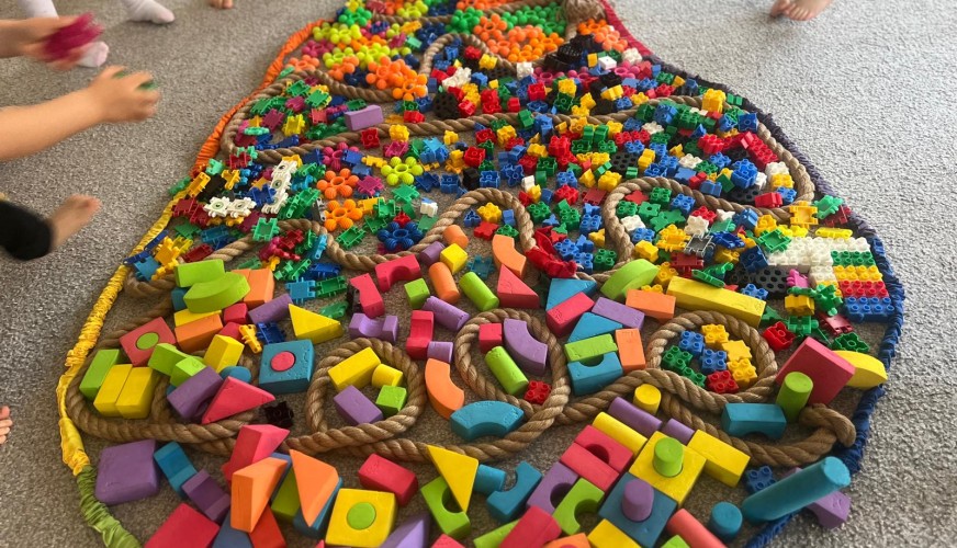 Duża dekoracyjna pisanka z kolorowych klocków Lego i drewnianych elementów, otoczona dziećmi w skarpetkach stojącymi na szary