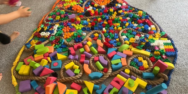 Duża dekoracyjna pisanka z kolorowych klocków Lego i drewnianych elementów, otoczona dziećmi w skarpetkach stojącymi na szary