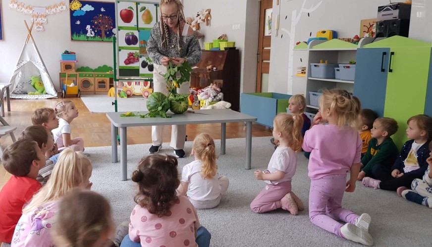 Nauczycielka pokazuje arbuza grupie około 15 dzieci siedzących na dywanie w sali przedszkolnej.