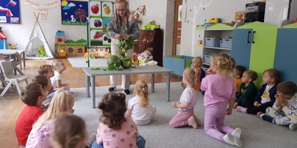 Nauczycielka pokazuje arbuza grupie około 15 dzieci siedzących na dywanie w sali przedszkolnej.
