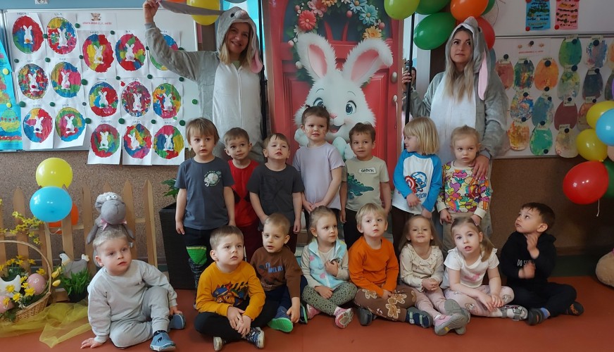 Grupa dzieci i dwóch opiekunów w przebraniach zajęczych w dekorowanej sali przedszkolnej z balonami, plakатami i jajkami