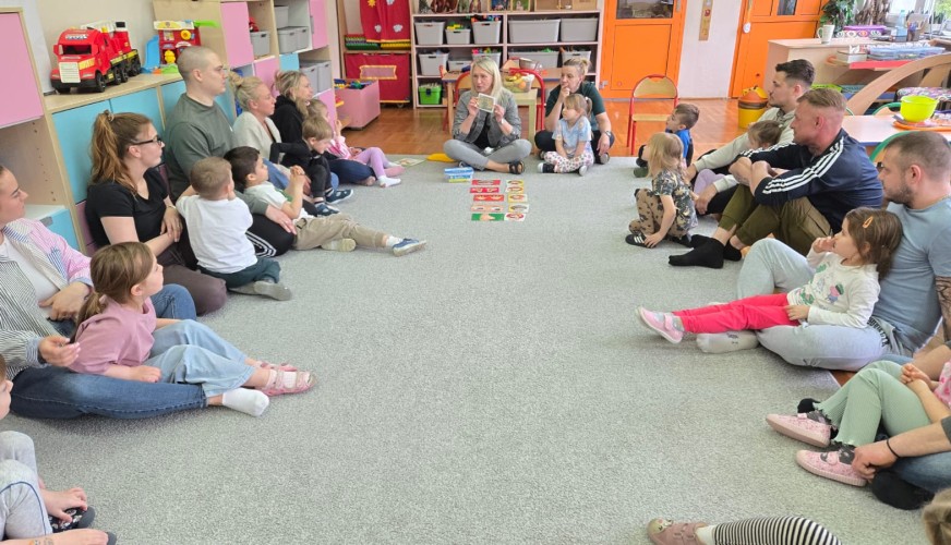 Grupa dzieci i dorosłych siedzi na podłodze w kole w kolorowej sali.