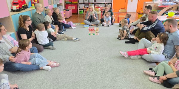 Grupa dzieci i dorosłych siedzi na podłodze w kole w kolorowej sali.