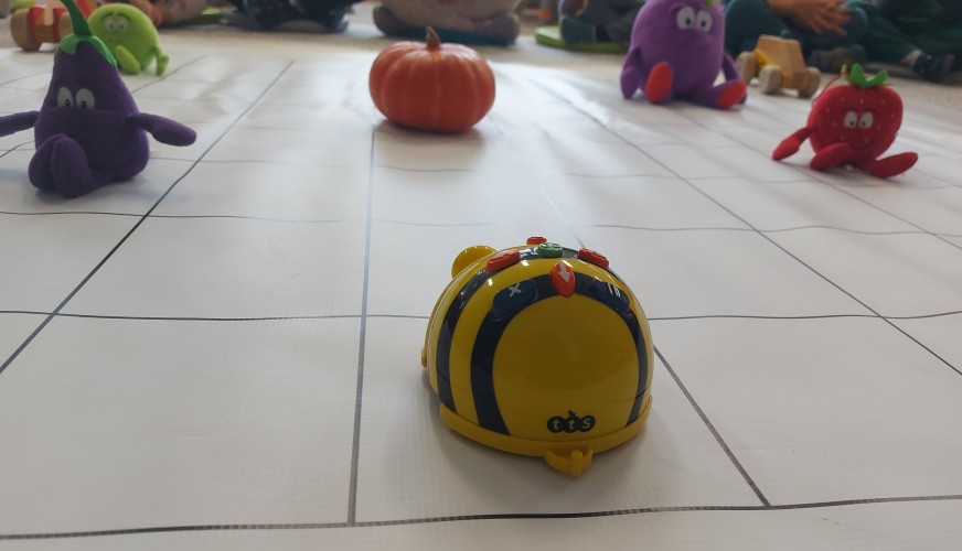 Robot Bee-Bot na białej macie z siatką, otoczony pluszowymi warzywami i dynią, przed grupą dzieci siedzących w kółku.