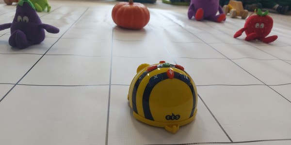 Robot Bee-Bot na białej macie z siatką, otoczony pluszowymi warzywami i dynią, przed grupą dzieci siedzących w kółku.