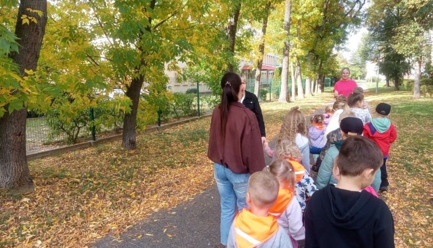 Grupa dzieci z przedszkola spaceruje alejką w parku jesienią, prowadzona przez dwie dorosłe opiekunki.