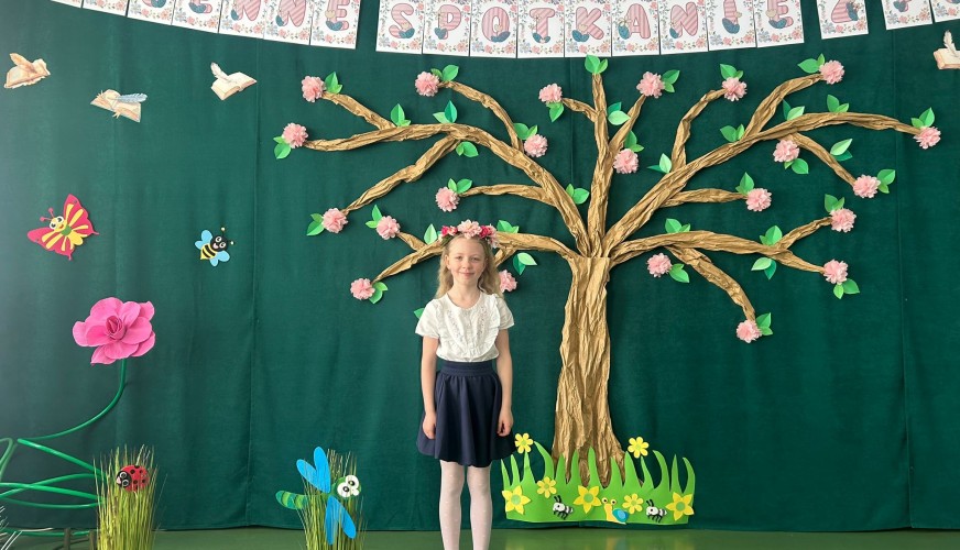 Dziewczynka stoi na zielonej scenie na tle dekoracji wiosennej z dużym złotym drzewem i różowymi kwiatami. Nad sceną wisi ban